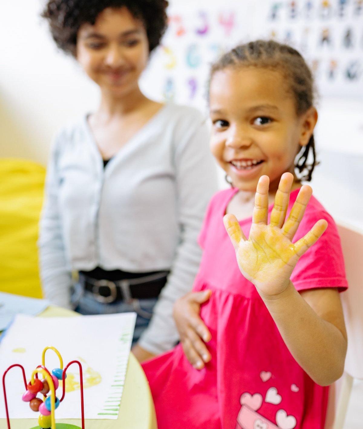 A smiling girl waving with a painted hand beside her teacher